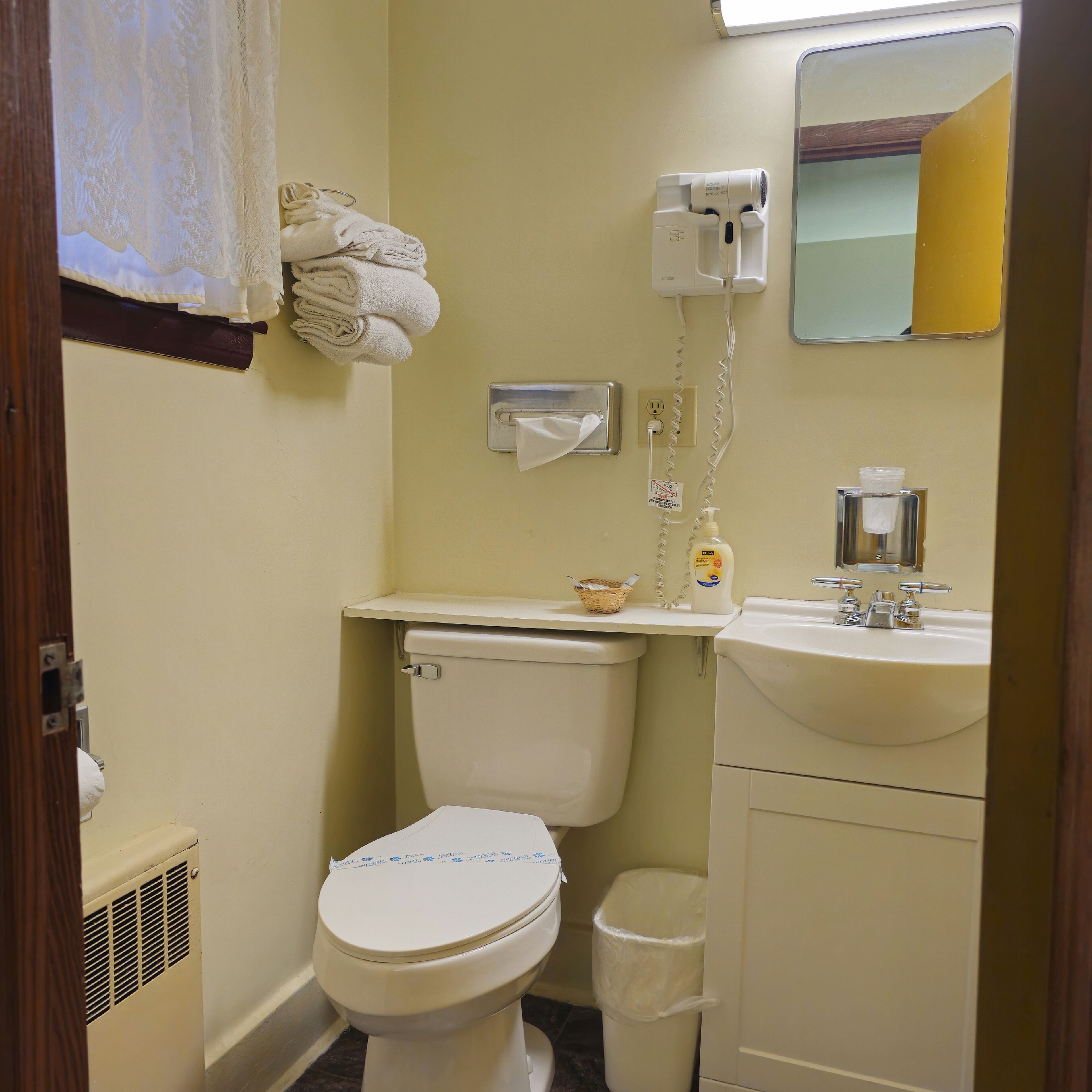 A small bathroom with a toilet, sink, and mirror. There are towels hanging on the wall and a trash can next to the toilet.
