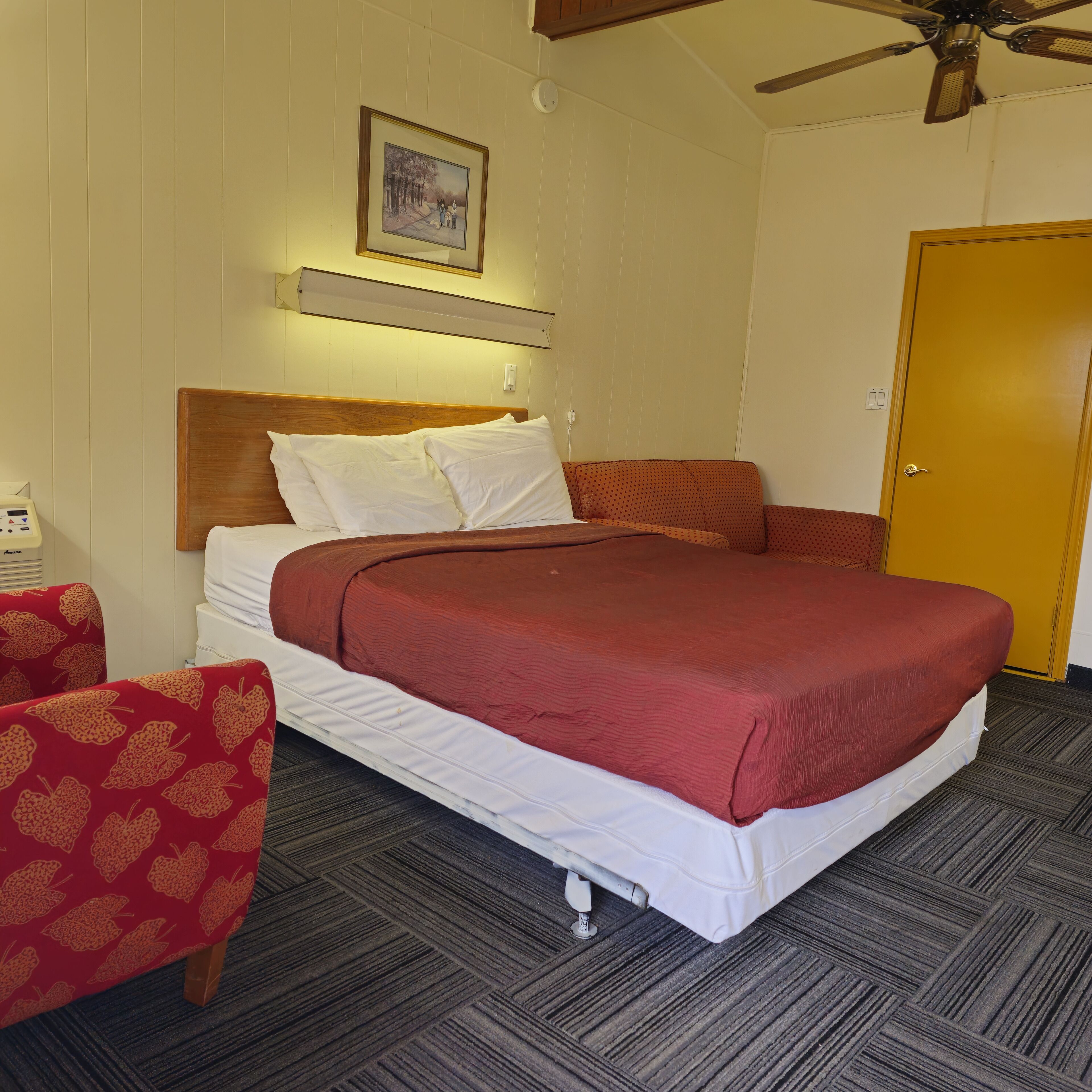 A hotel room with a bed, a wooden headboard, a ceiling fan, a framed picture on the wall, and a door.