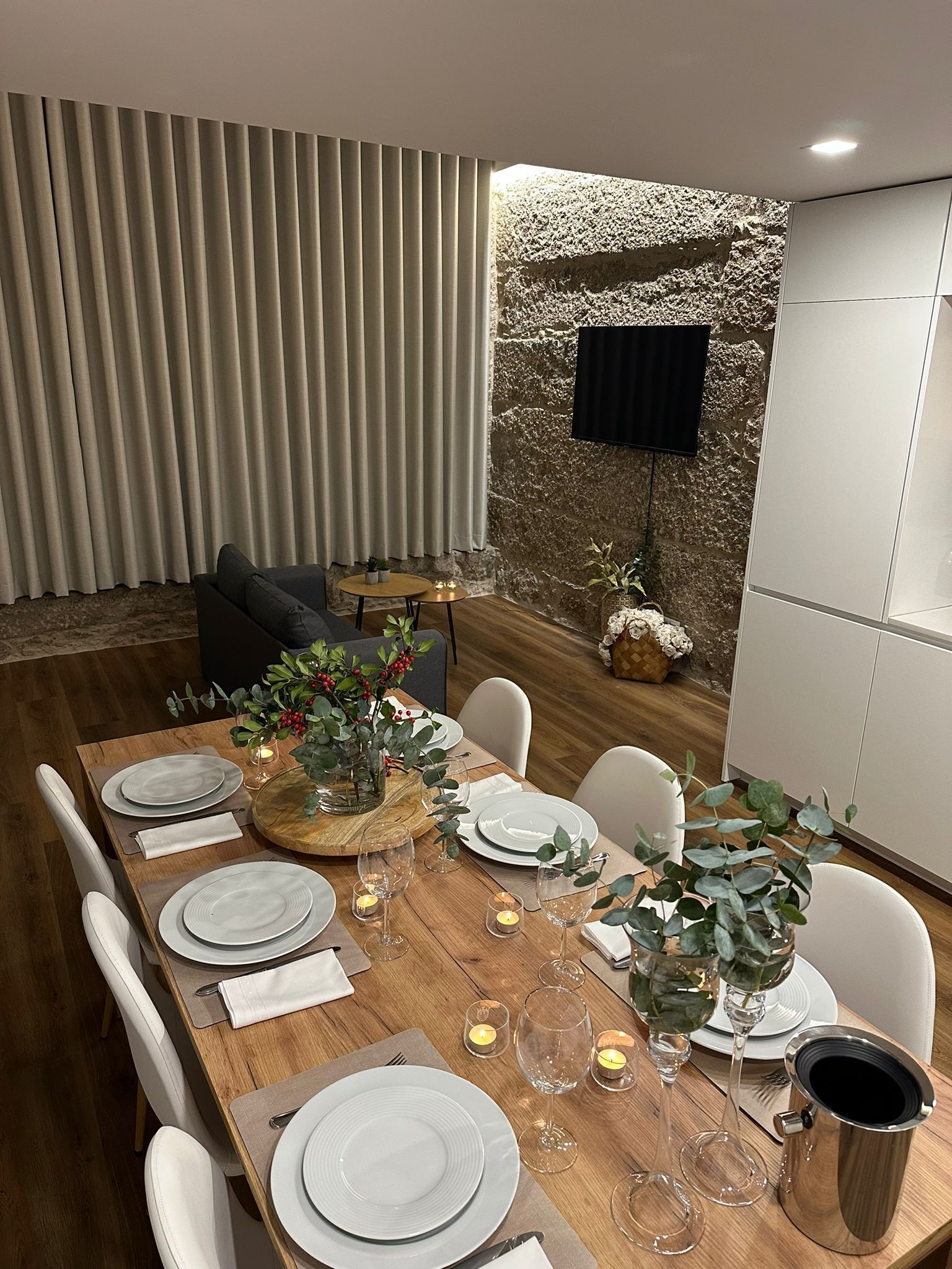 Un coin repas avec une table en bois dressée pour six personnes, entourée de chaises blanches. On aperçoit un mur en pierre avec un téléviseur fixé au mur à l’arrière-plan.