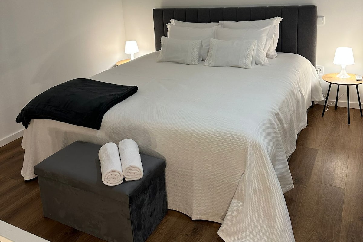 Une chambre d’hôtel moderne avec un grand lit, deux tables de chevet avec des lampes, un petit banc avec des serviettes et un plancher en bois.