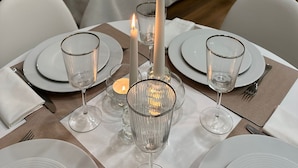 Una mesa de comedor elegantemente dispuesta con velas, vasos y platos en una habitación de hotel moderna con un muro de acento de piedra y una cama al fondo.