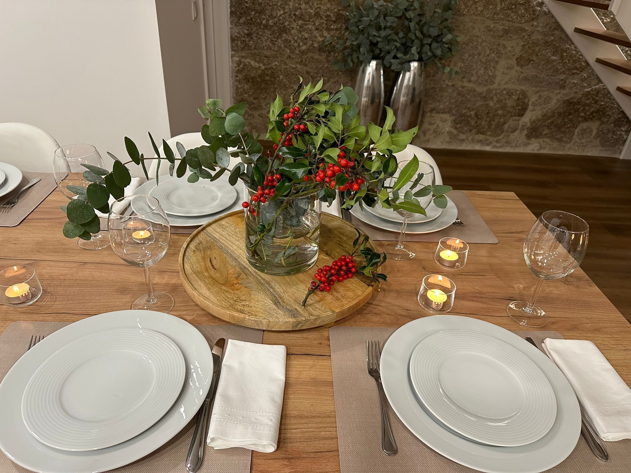 A dining table set with white plates, cutlery, and a centerpiece of red berries in a glass vase.