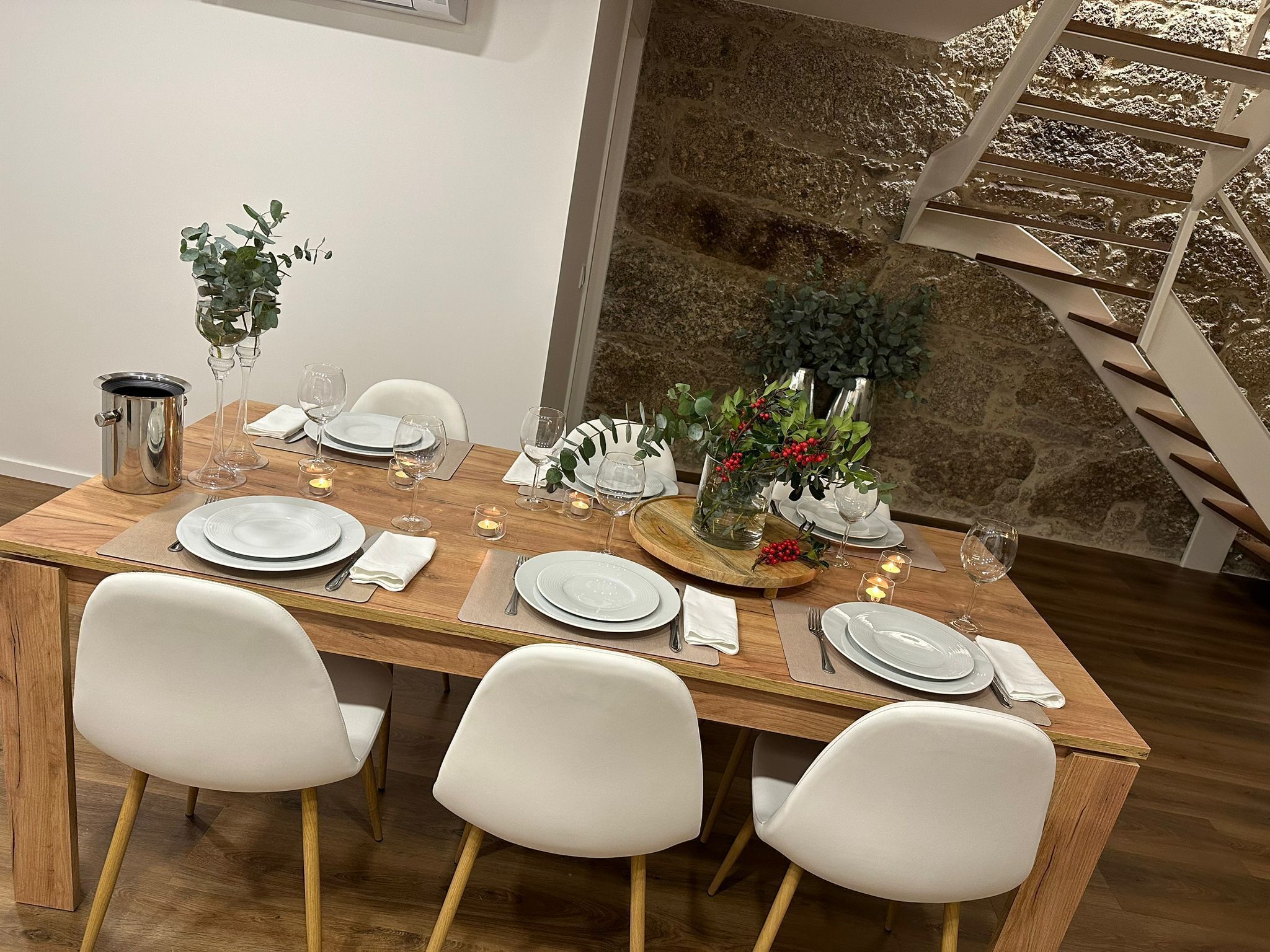 Una mesa de comedor dispuesta con platos blancos, vasos y cubiertos, rodeada de sillas blancas.