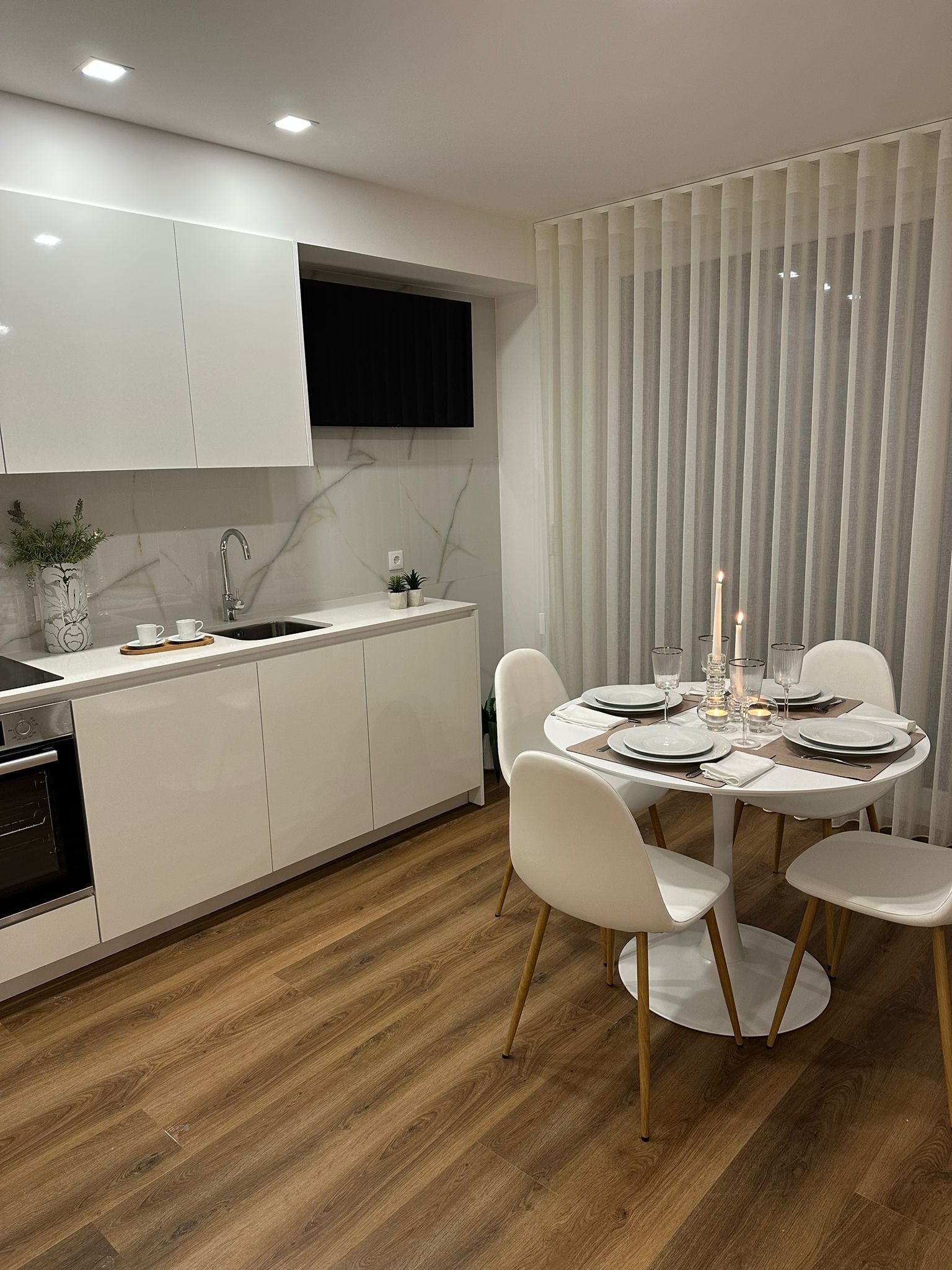 Een moderne keuken met witte kasten, een inbouwoven en een eethoek met een ronde tafel gedekt voor vier personen.