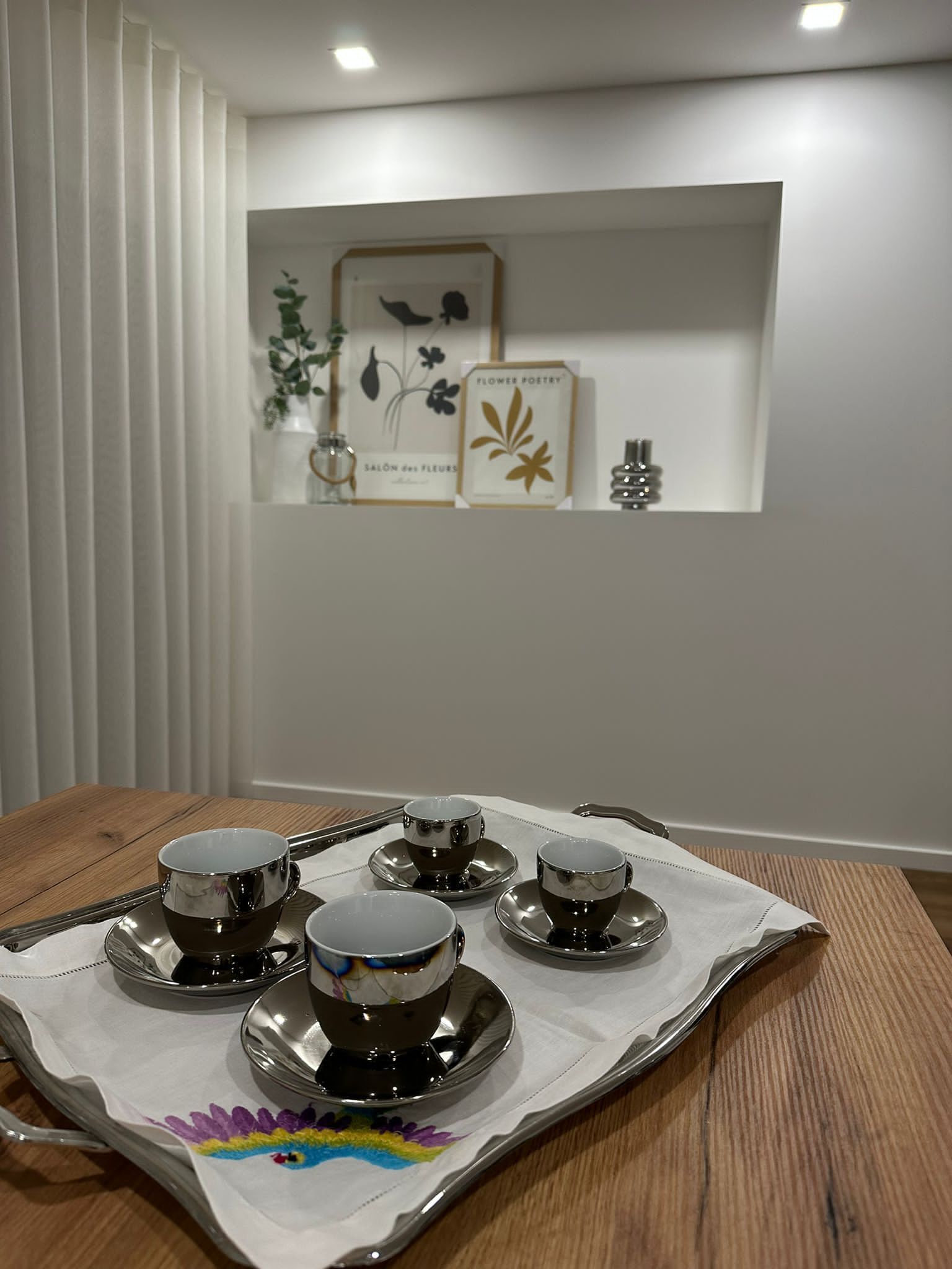 A dining table with a tray holding four cups and saucers, a decorative cloth with a colorful design, and a wall with framed artwork and a small plant.