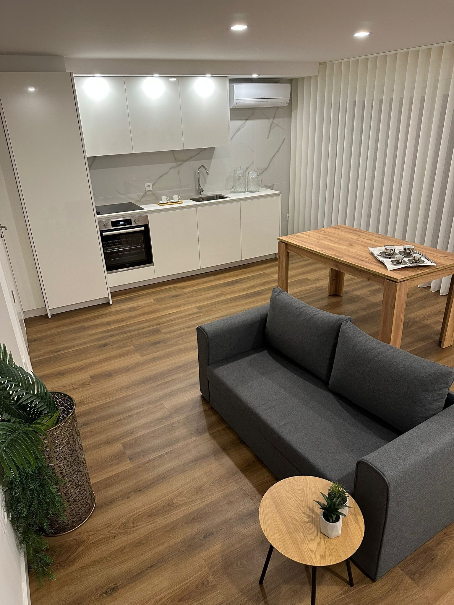 Uma sala de estar moderna com um sofá cinzento, uma mesa de jantar de madeira e uma pequena mesa redonda com uma planta. Uma cozinha com armários brancos e um forno é visível ao fundo.