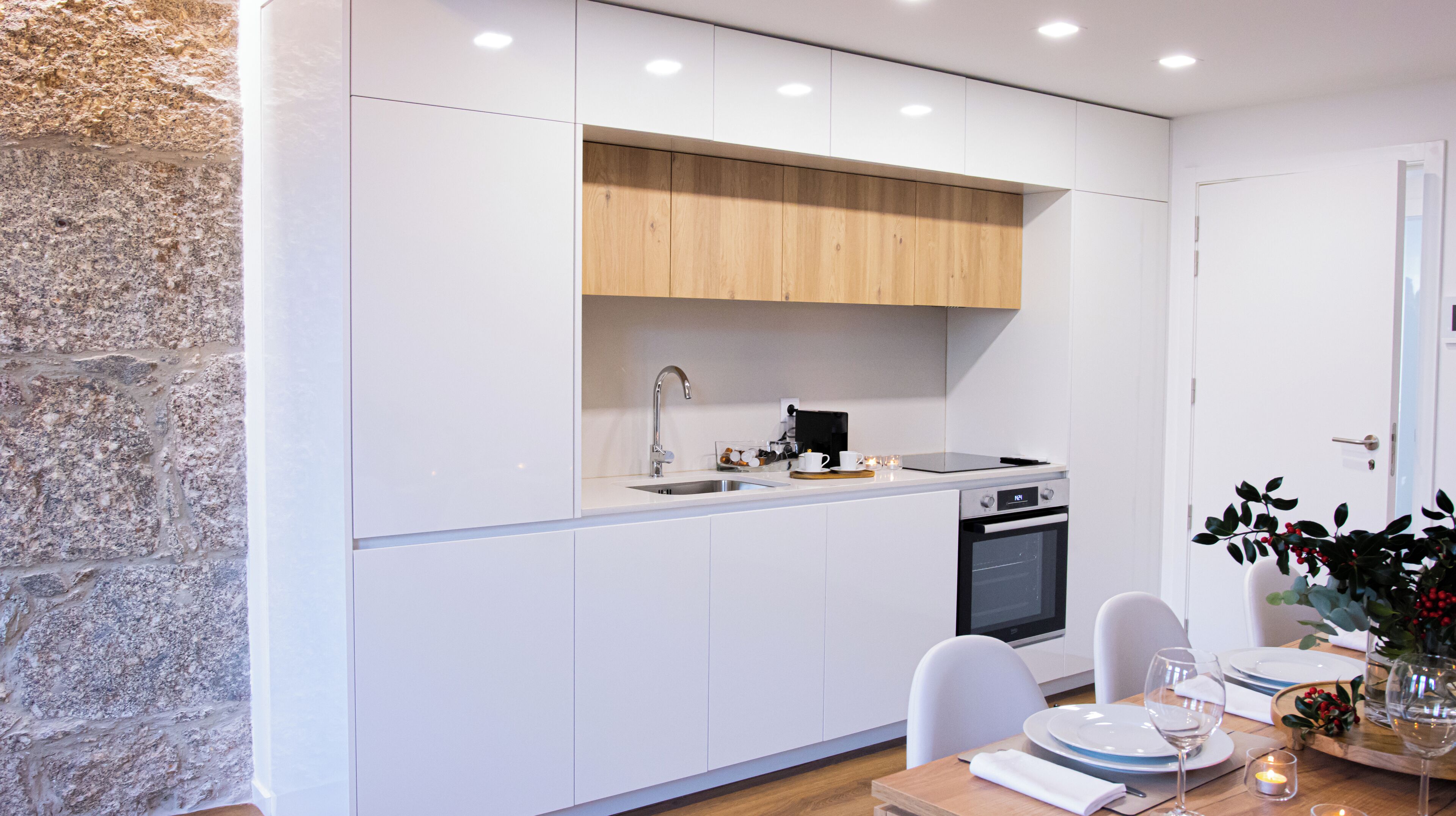Une cuisine moderne avec des armoires blanches, un plan de travail en bois, un four encastré et un coin repas avec une table mise pour deux personnes.