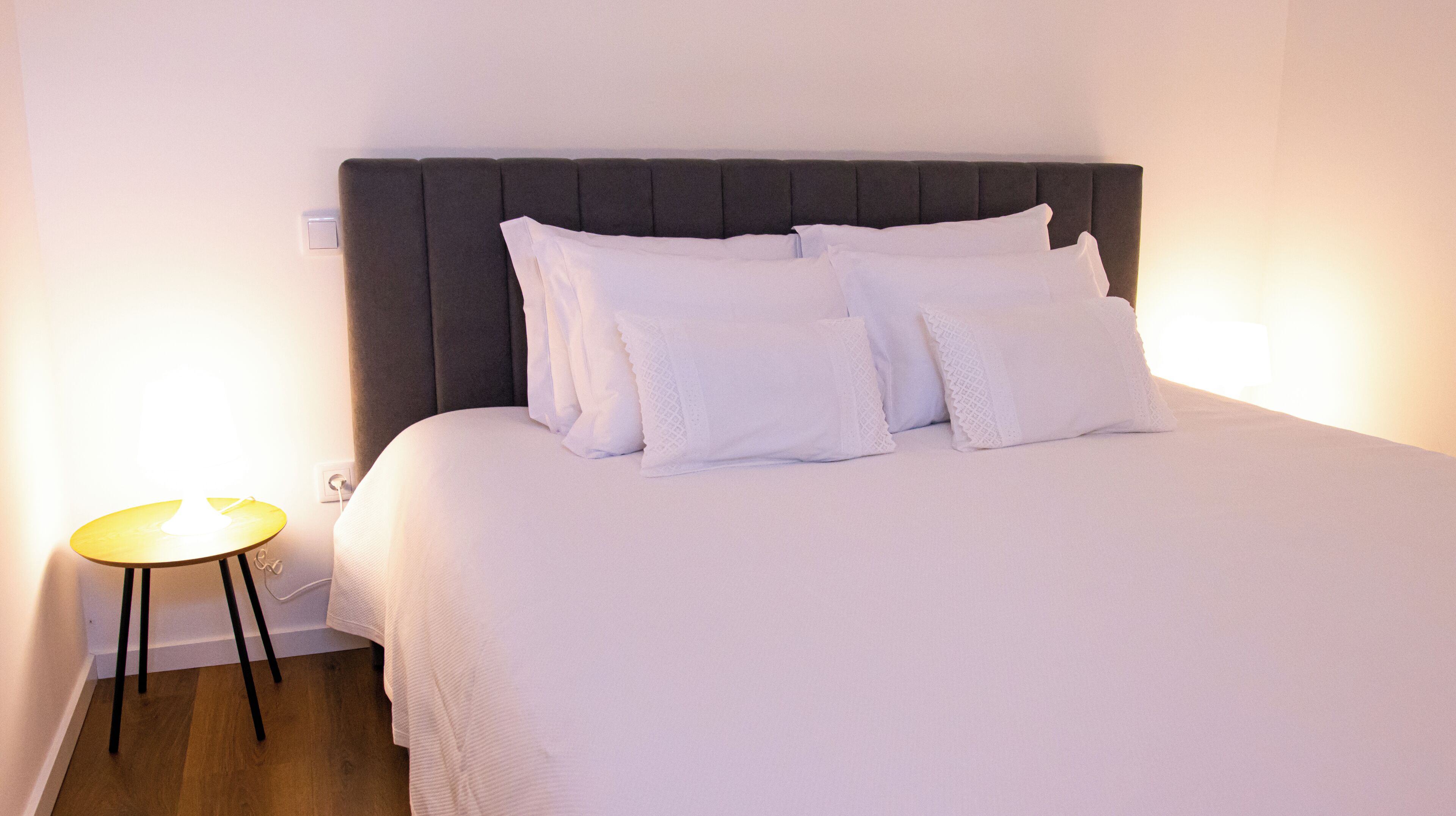 A neatly made bed with white linens and pillows, a dark headboard, and a small table with a lamp beside it.