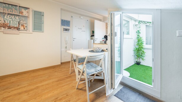 Een compacte keuken met een eettafel en stoelen, een witte koelkast, en een klein raam met uitzicht op een groen buitengebied.
