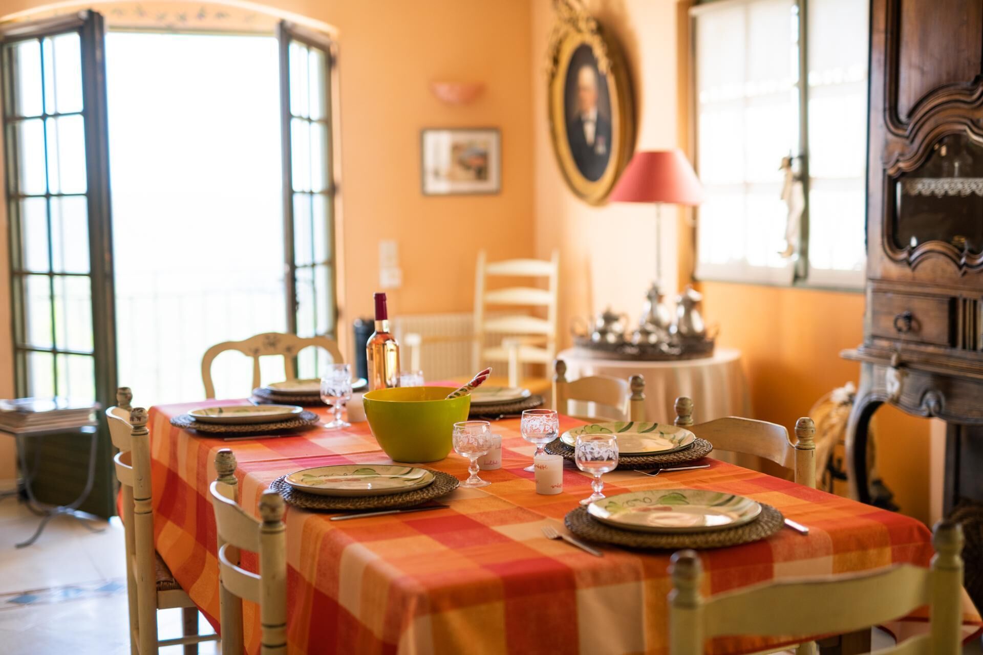 A dining table set with plates, glasses, and a bowl, surrounded by chairs in a warmly lit room with a vintage aesthetic.