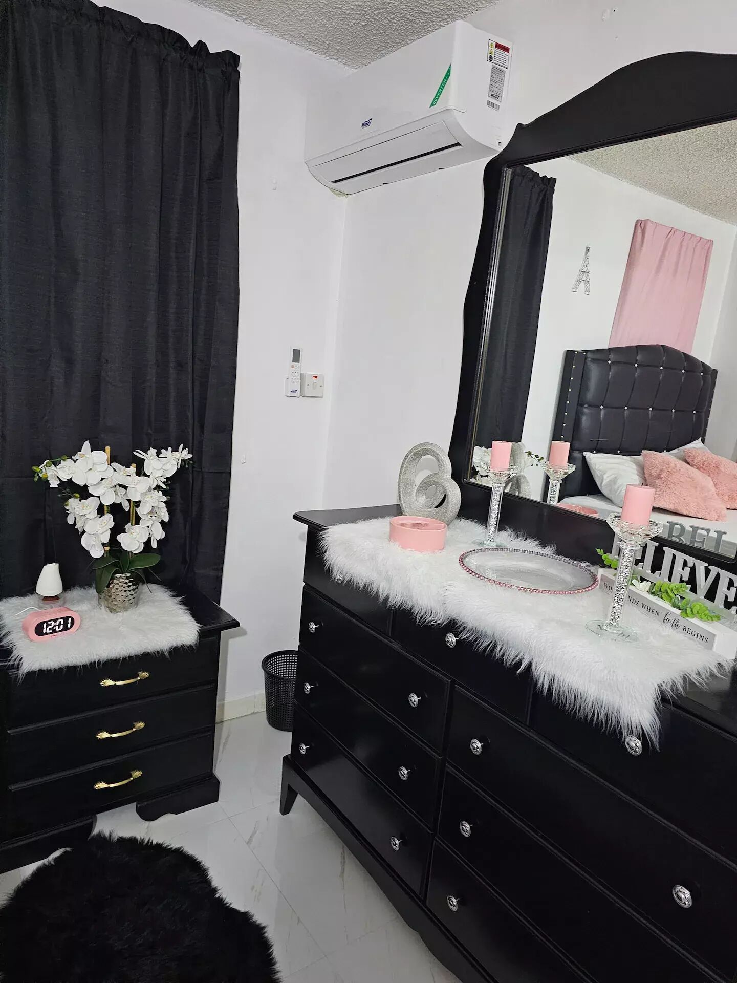 Une chambre avec une commode noire, un miroir, une coiffeuse avec une bougie rose et un cadre de miroir décoratif représentant la Tour Eiffel.