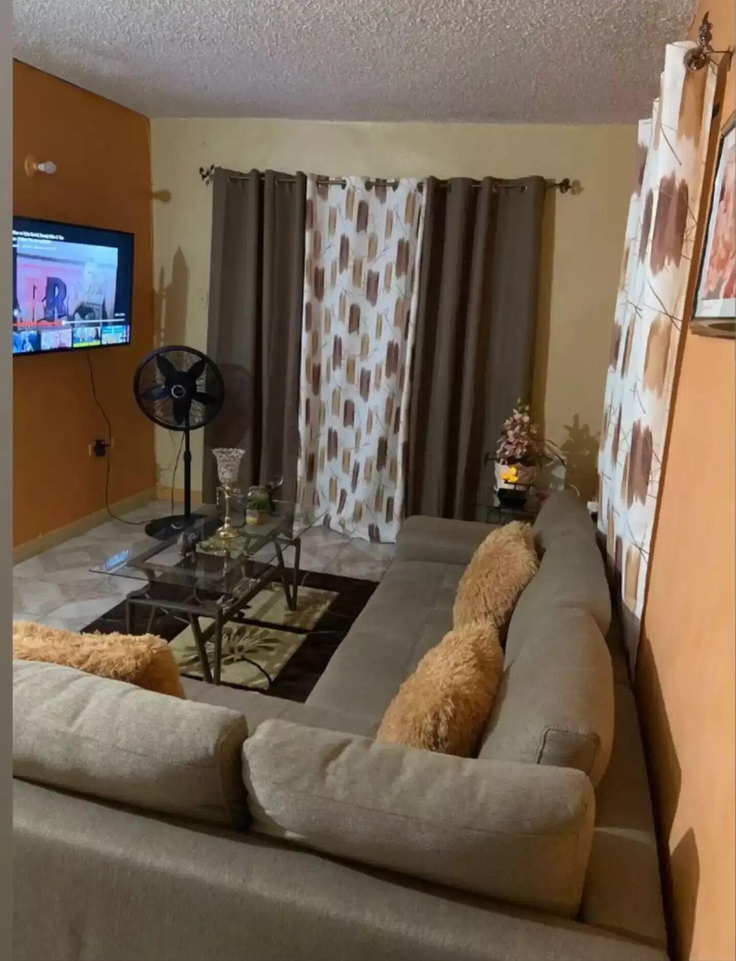 A living room with a sofa, a glass coffee table, a fan, and a TV on the wall.