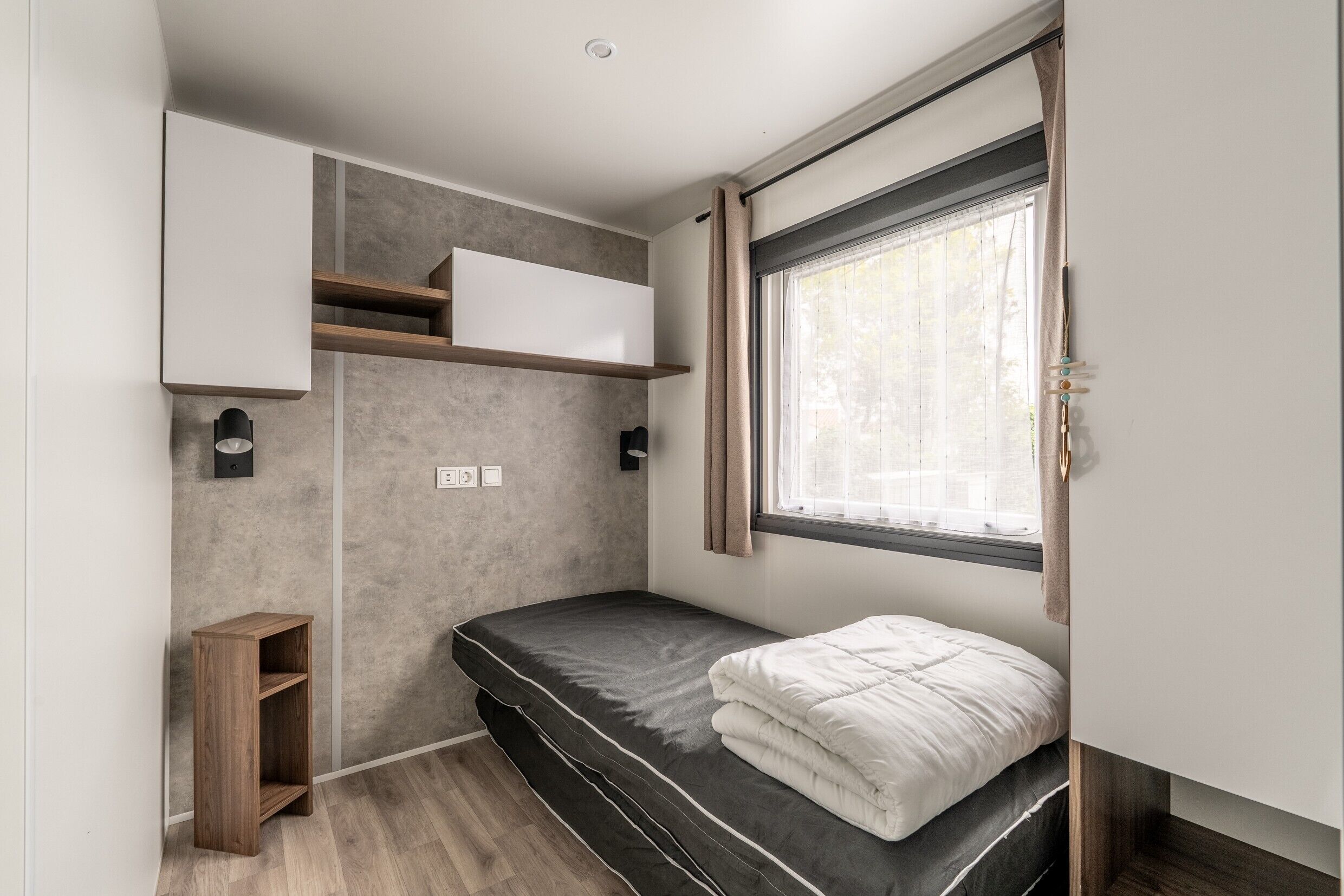 Une petite pièce comprenant un lit, une petite étagère en bois, une lampe fixée au mur et une fenêtre avec des rideaux.