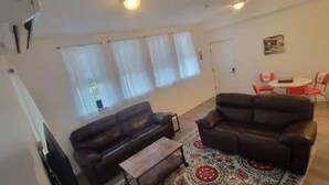 A living room with a sofa, a coffee table, and a patterned rug. There is a window with curtains, a door, and a small table with chairs. - Historic Chesapeake City Stay   (Chesapeake City)