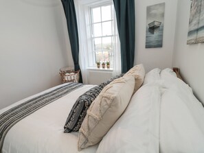 A neatly made bed with a striped blanket and patterned pillows, a window with curtains, and a small table with potted plants. - Victoria House (Belford)