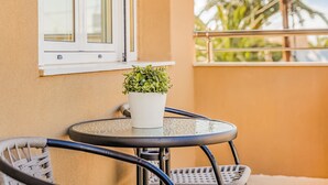 A small balcony with a round table and two wicker chairs, a potted plant, and a window with a view of palm trees. - Maela Luxury Suite (Hersonissos)