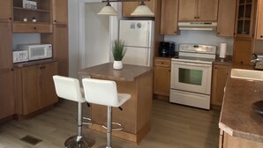 A modern kitchen with wooden cabinets, a central island, two white bar stools, a refrigerator, a stove, and a microwave. - Cape May Home in Newest Hot Spot: Centrally Located, Family Friendly, Sleeps 12! (Cape May)