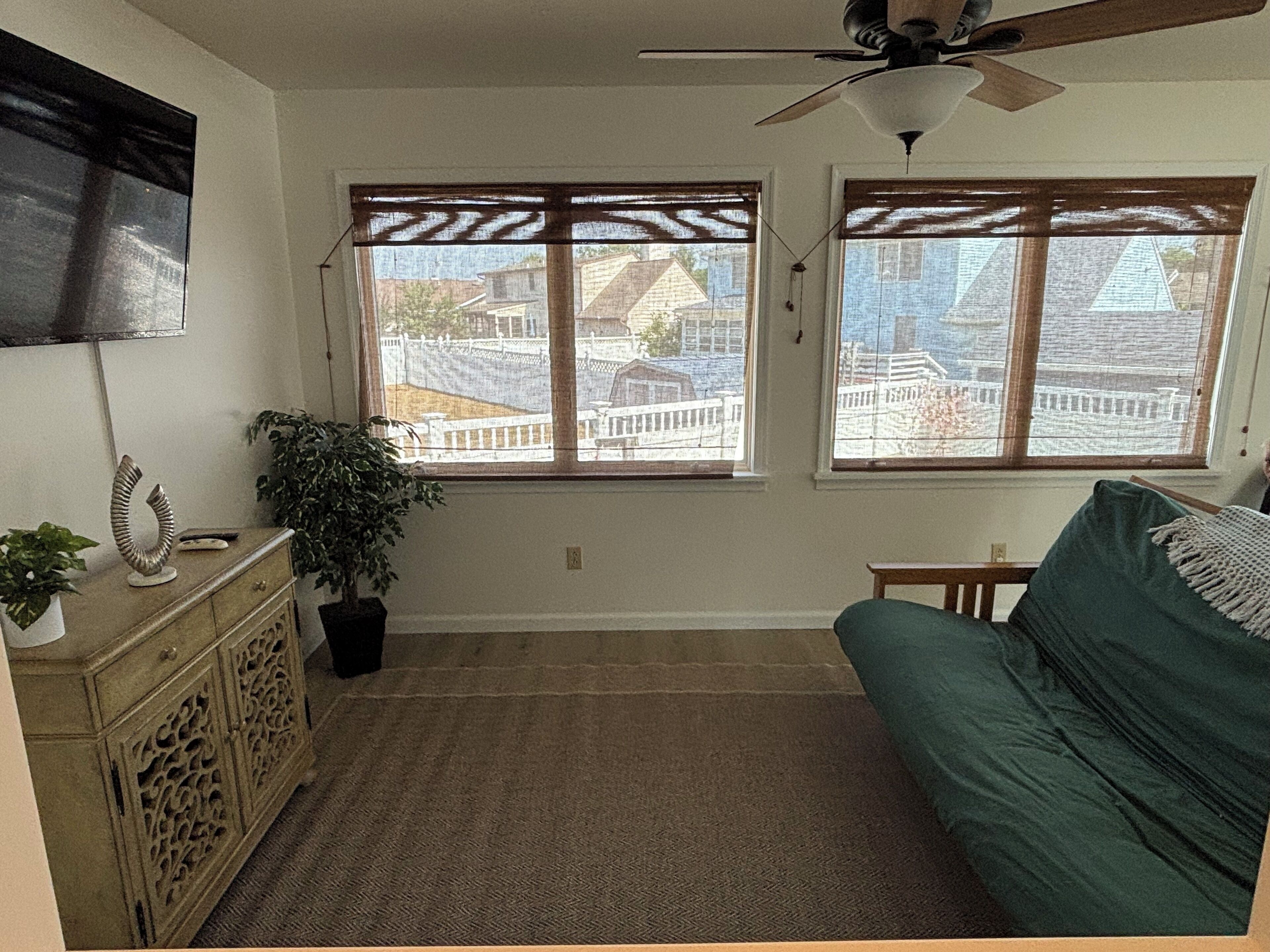 Una habitación con ventilador de techo, dos ventanas con persianas, un sofá verde, un armario de madera y una planta en maceta.