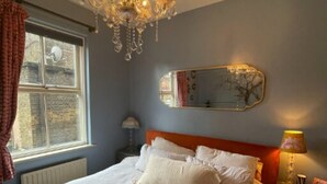 Un dormitorio con una cama grande, un candelabro, una ventana con cortinas, una mesita de noche con una lámpara y un espejo en la pared.