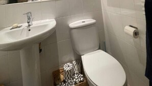Un baño con un lavamanos blanco, inodoro y un espejo sobre el lavamanos. Hay artículos de aseo en el lavamanos y un perchero encima del inodoro.
