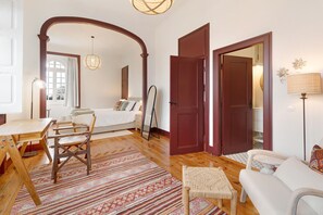 Um quarto com cama, uma mesa de madeira, cadeira de fibra natural, tapete estampado e sofá branco.