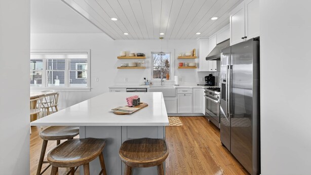 Una cocina moderna con armarios blancos, una isla central con taburetes, un refrigerador de acero inoxidable y suelo de madera.