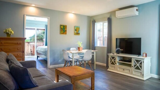 A living room with a sofa, a coffee table, a TV on a white cabinet, and a dining area with a table and chairs. - Relaxing Home With SF Bay View

 (Millbrae)