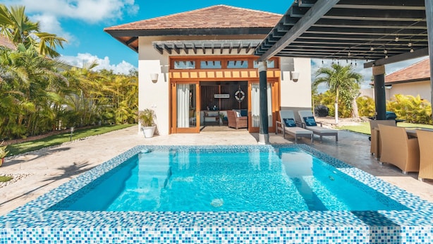 A poolside area with a clear blue pool, a covered seating area, and a building with a tiled roof. - Bungallow With Private Backyard and Pool (Punta Cana)