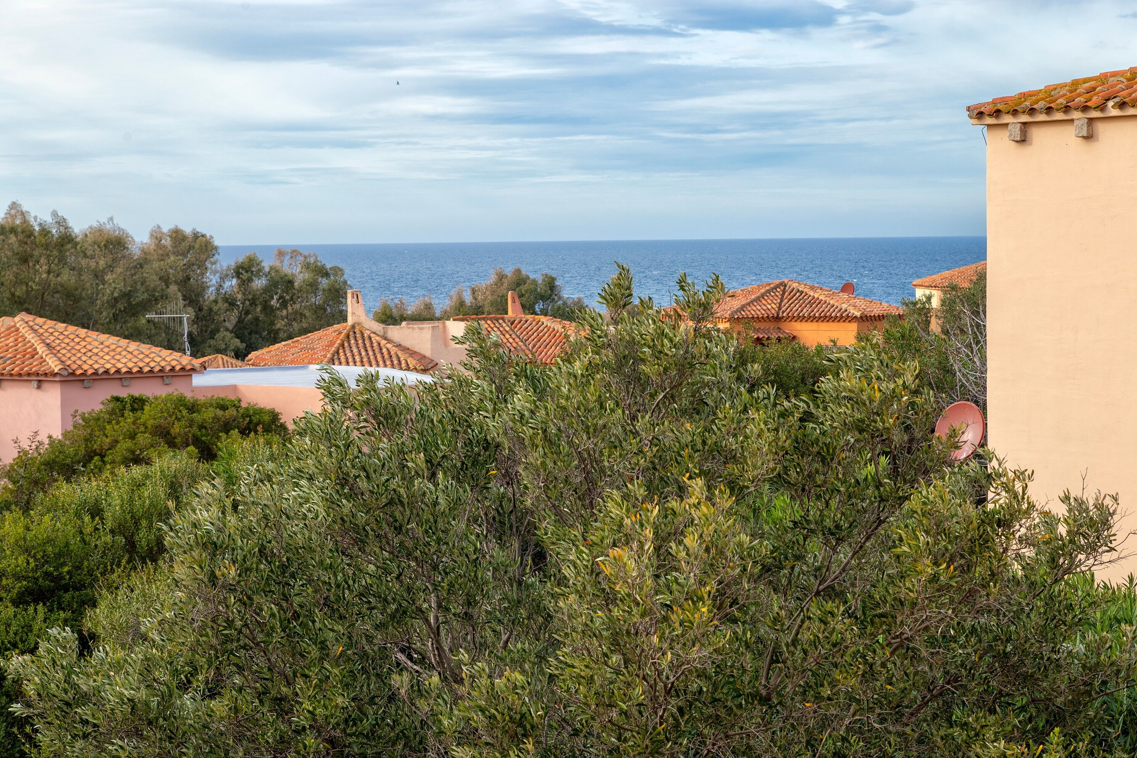 Una zona residencial costera con edificios de tejados de terracota, abundante vegetación y vista al mar.