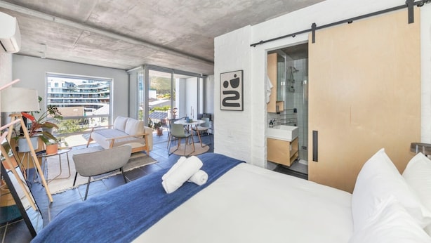 A modern hotel room with a large bed, a blue throw blanket, and a view of the city through large windows.