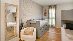Ein Schlafzimmer mit weißer Backsteinwand, einem Bett mit grauem Kopfteil, einem weißen Stuhl und einem Spiegel mit Holzrahmen.