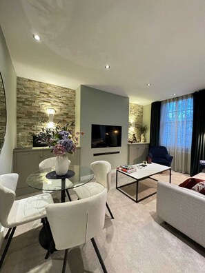 Un salon moderne avec une table à manger en verre, des chaises blanches, un canapé, une table basse et un téléviseur à écran plat fixé au mur.