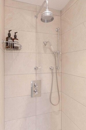 Un espace douche équipé d’un pommeau de douche à main, d’un robinet mural et d’un panneau de commande. On y trouve une étagère avec deux flacons de produits de douche de la marque Aesop.