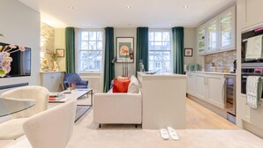 A modern living room with a sofa, a glass coffee table, and a TV. There is a kitchen area with cabinets and appliances. - Elegant 1BD Flat in the Heart of Notting Hill! (London)