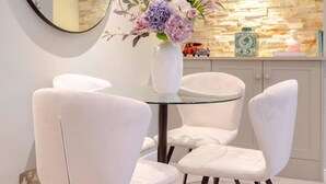 A dining area with a round glass table, four white upholstered chairs, a vase with flowers, a stone wall, and a mirror on the wall. - Elegant 1BD Flat in the Heart of Notting Hill! (London)