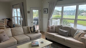 A living room with two beige sofas, a coffee table, and a view of a green landscape through large windows. - Renovated, Golf cart included, pet friendly, club cards available - 326 Ocean Point (Fripp Island)