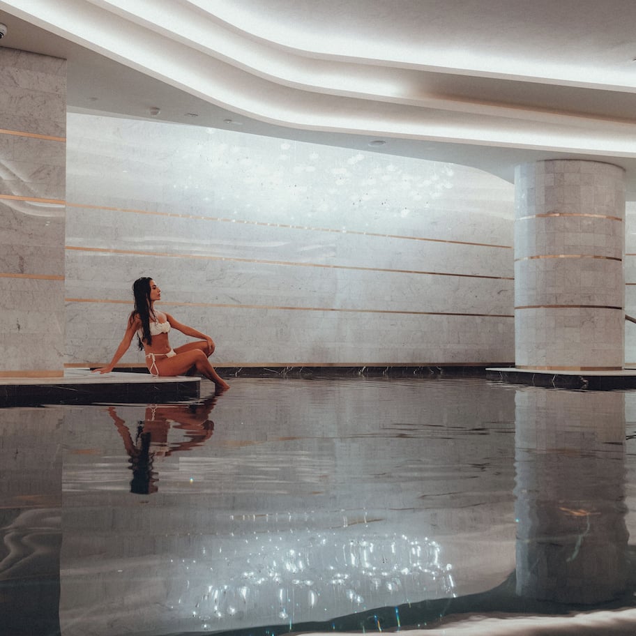 A spacious indoor pool area with a marble floor and walls, a person sitting by the pool, and a modern architectural design.