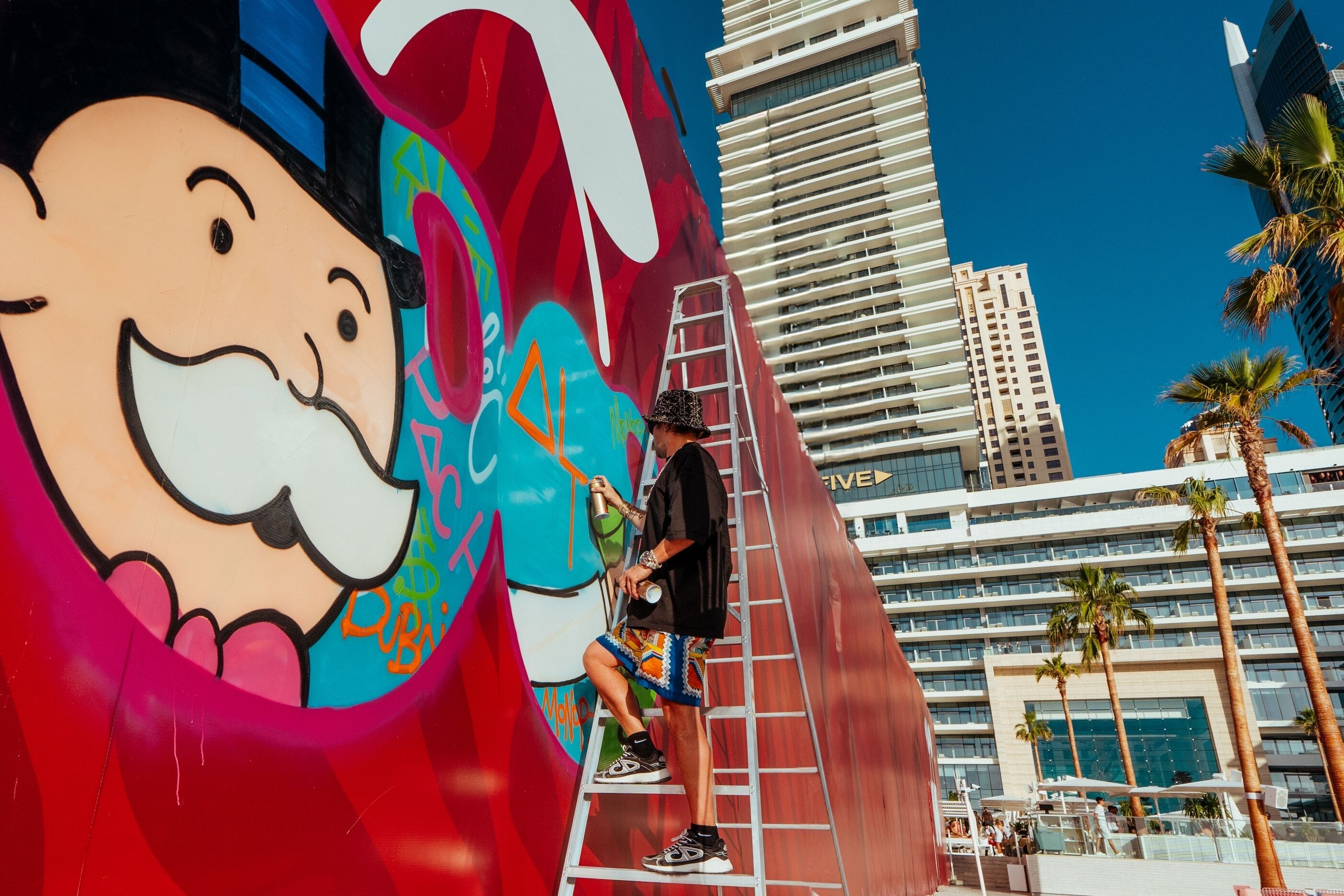 Un murale raffigura un personaggio dei cartoni animati, una scala e un edificio sullo sfondo con palme.