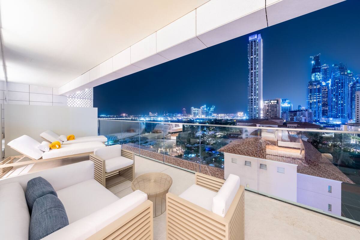 Un balcón moderno y bien iluminado con vistas a la ciudad de noche, que presenta mobiliario contemporáneo y barandilla de vidrio.