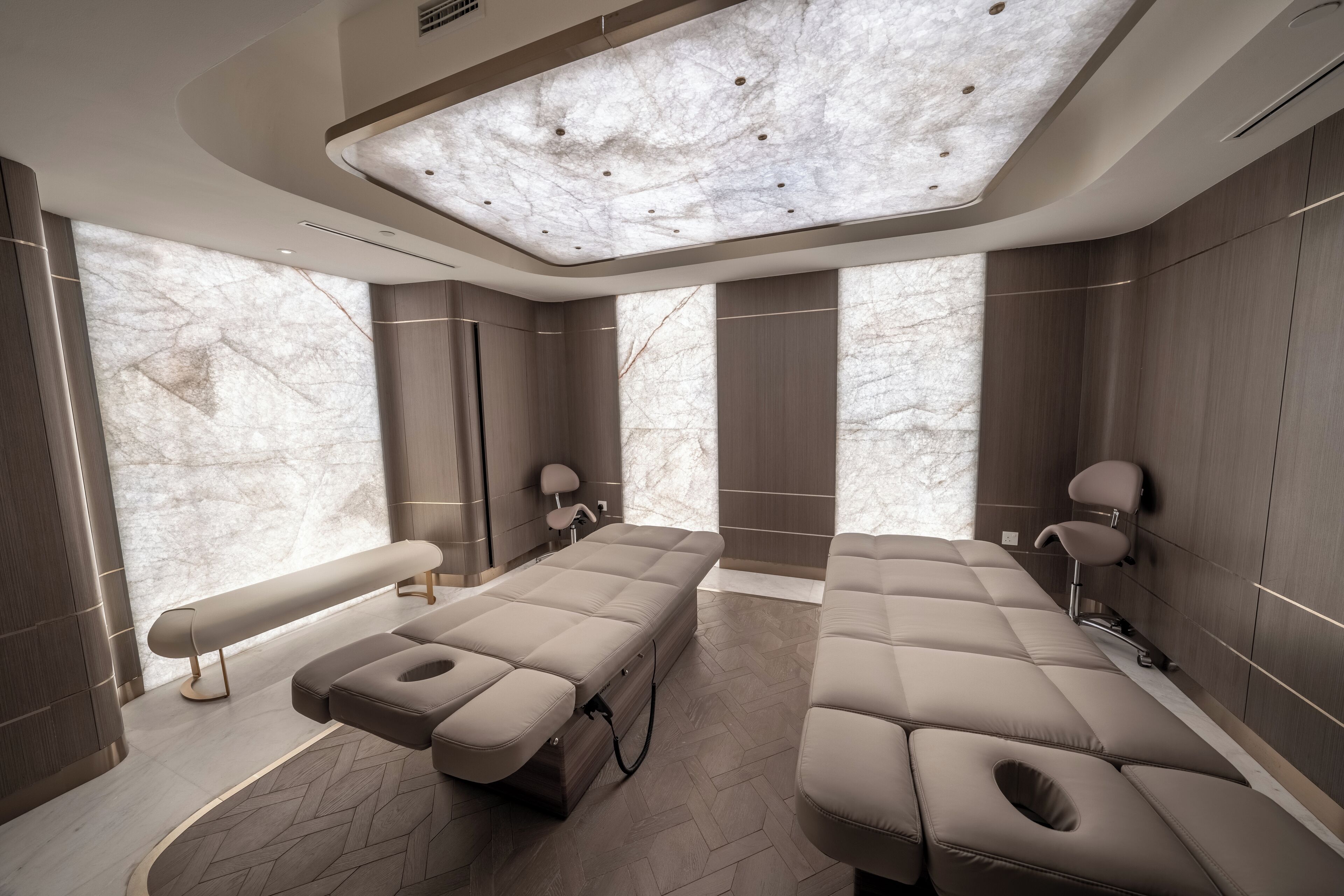 A modern spa room with a large massage table, a smaller massage table, and a bench.