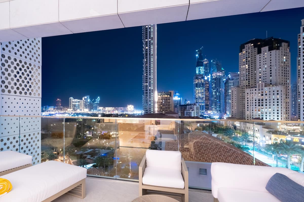 Un balcón moderno con mobiliario blanco, barandilla de vidrio y vista nocturna de la ciudad.