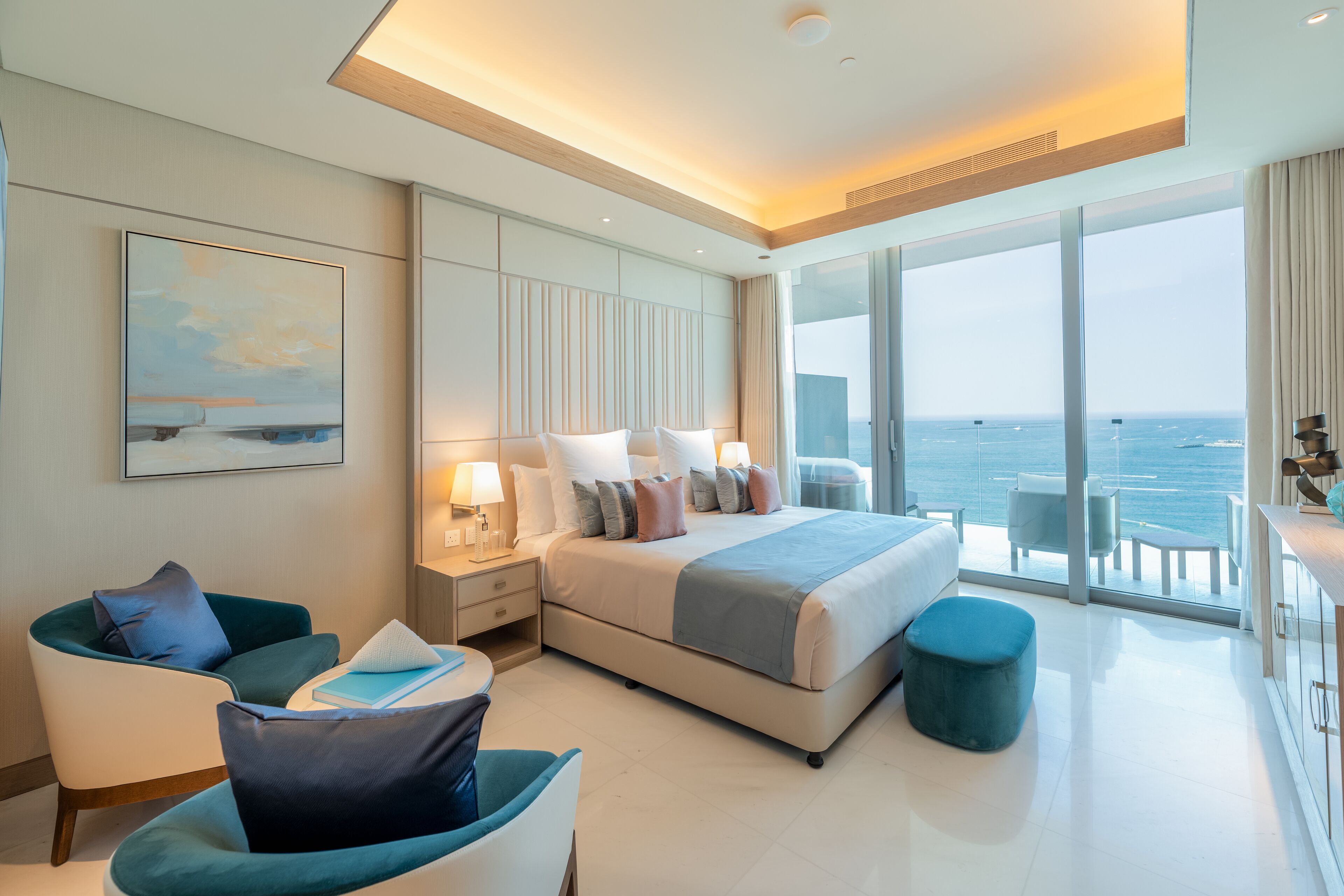 A modern hotel room with a large bed, a seating area with a sofa and armchair, a dining area, and a view of the ocean.