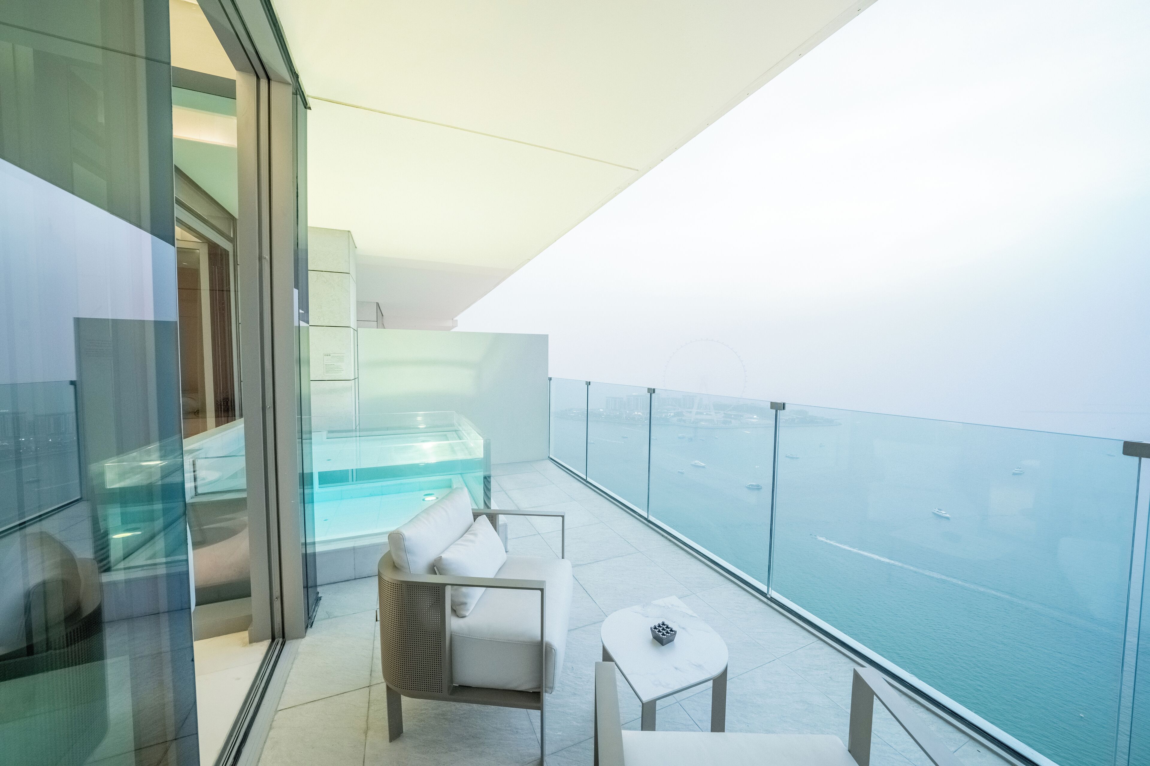 A modern balcony with a glass railing, a white sofa, a small table, and a glass-enclosed hot tub.