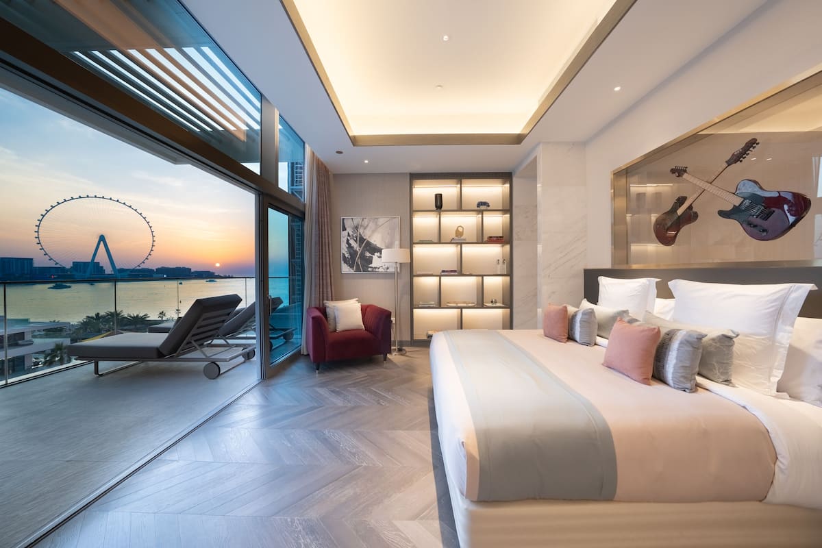 Una habitación de hotel moderna con una cama grande, un sillón rojo, un muro de cristal con vistas a una noria y una guitarra en la pared.