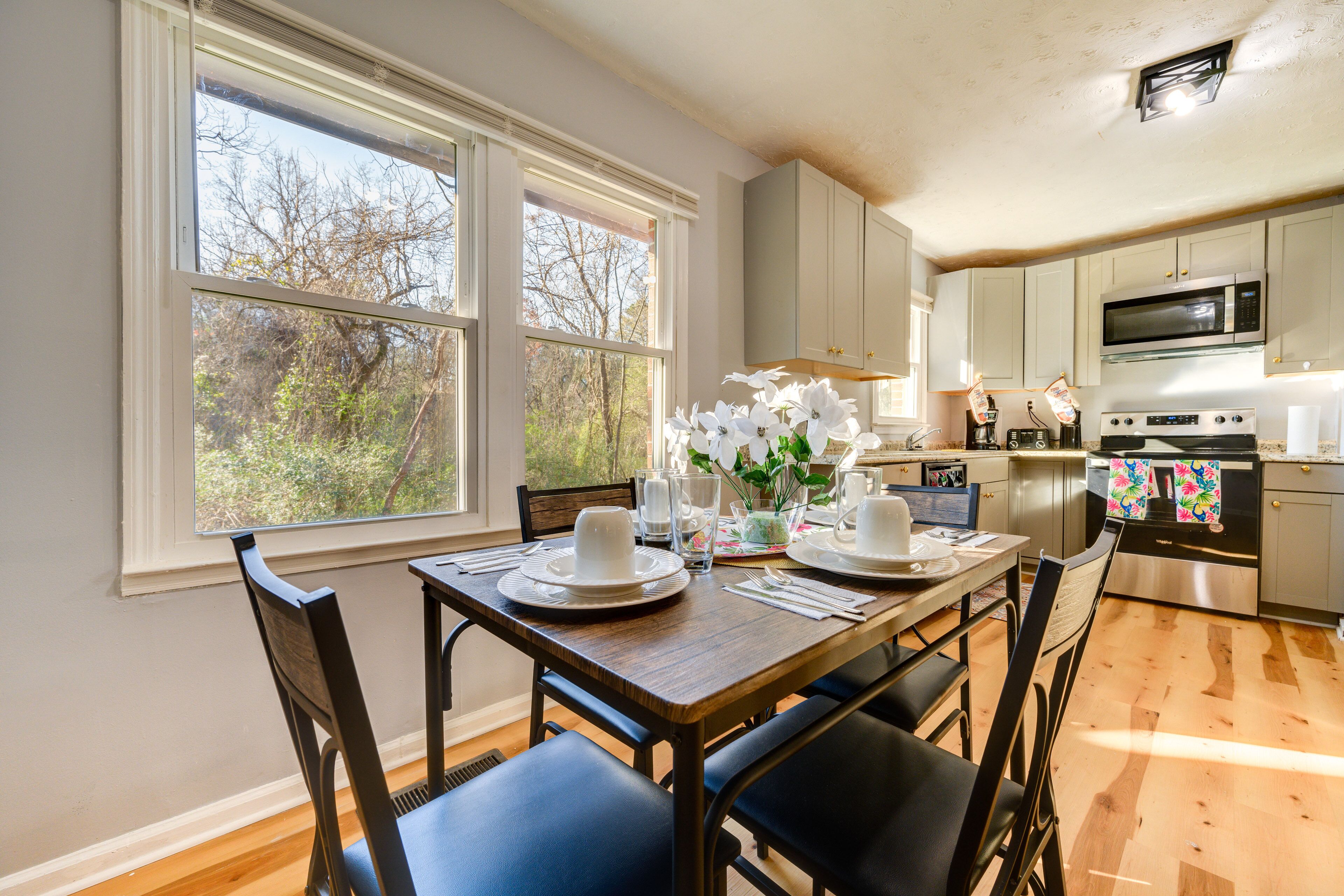 Un coin repas avec une table en bois dressée pour quatre personnes, une cuisine équipée d’appareils modernes et de grandes fenêtres offrant une vue sur les arbres.