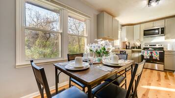Un coin repas avec une table en bois dressée pour quatre personnes, une cuisine équipée d’appareils modernes et de grandes fenêtres offrant une vue sur les arbres.