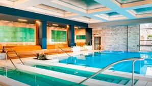 Une piscine intérieure moderne, dotée d’une eau claire, de sièges en bois et d’un mur en pierre.