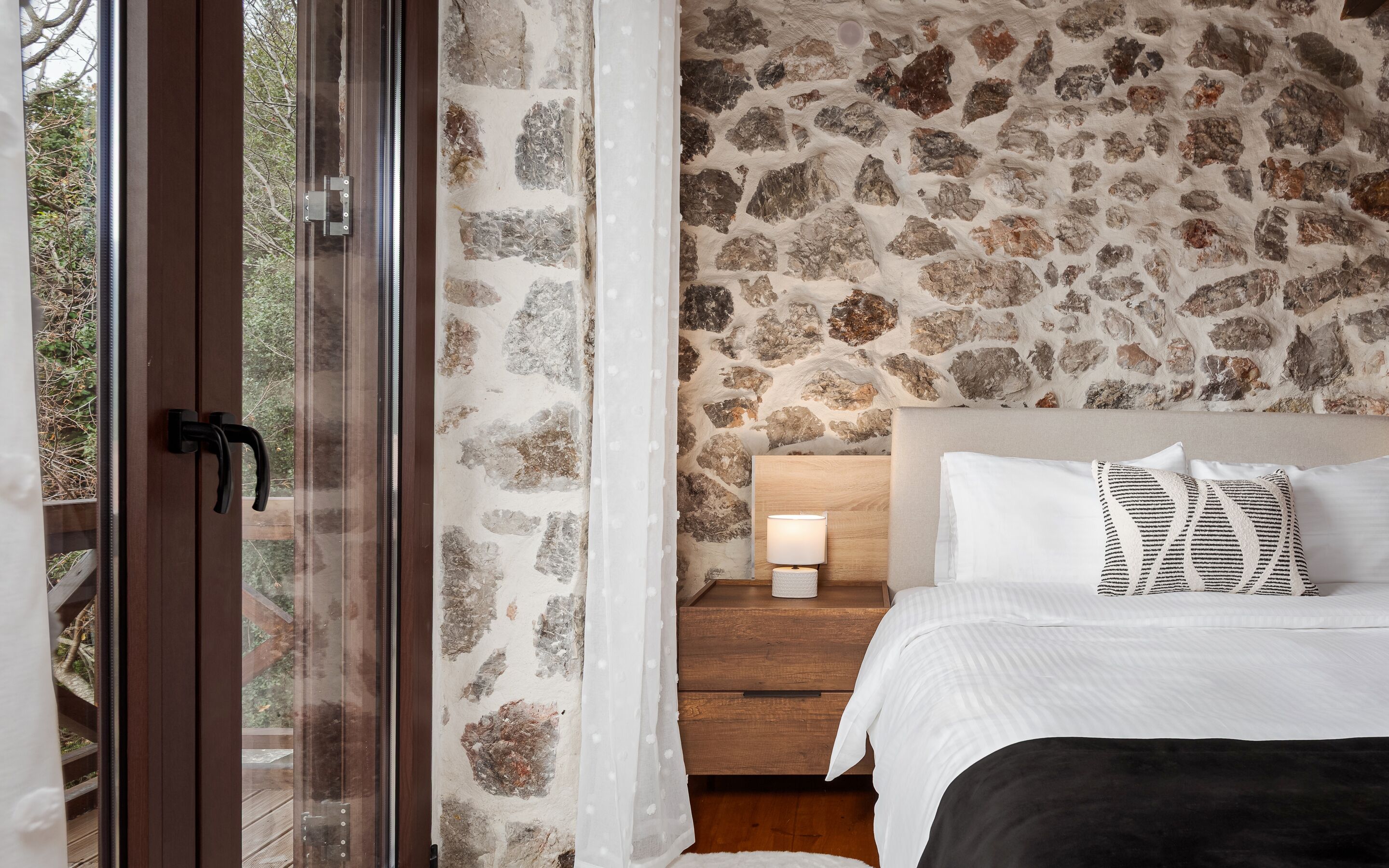 A bedroom with a stone wall, a wooden nightstand with a lamp, and a bed with white bedding and patterned pillows.