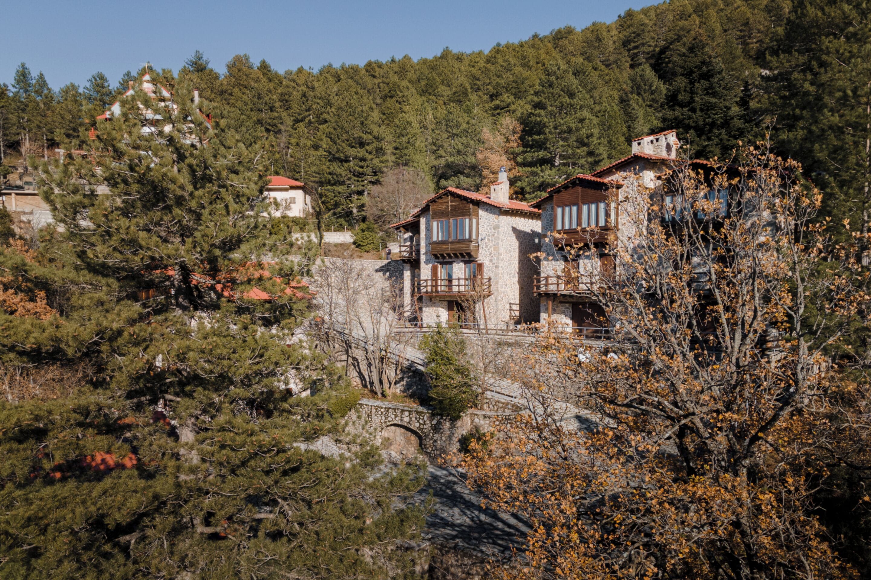 Um edifício de pedra com várias varandas e telhado de telhas vermelhas, rodeado por árvores e uma paisagem montanhosa ao fundo.