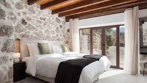 A bedroom with a stone wall, wooden ceiling beams, a large bed with white bedding, and a view through glass doors. - Parnassus Woodstone Chalets- N1- N2- N3 (Delphi)